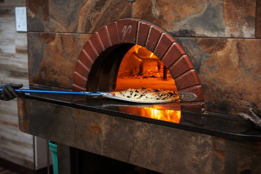 A pizza is being placed into a wood-fired oven with a metal peel.