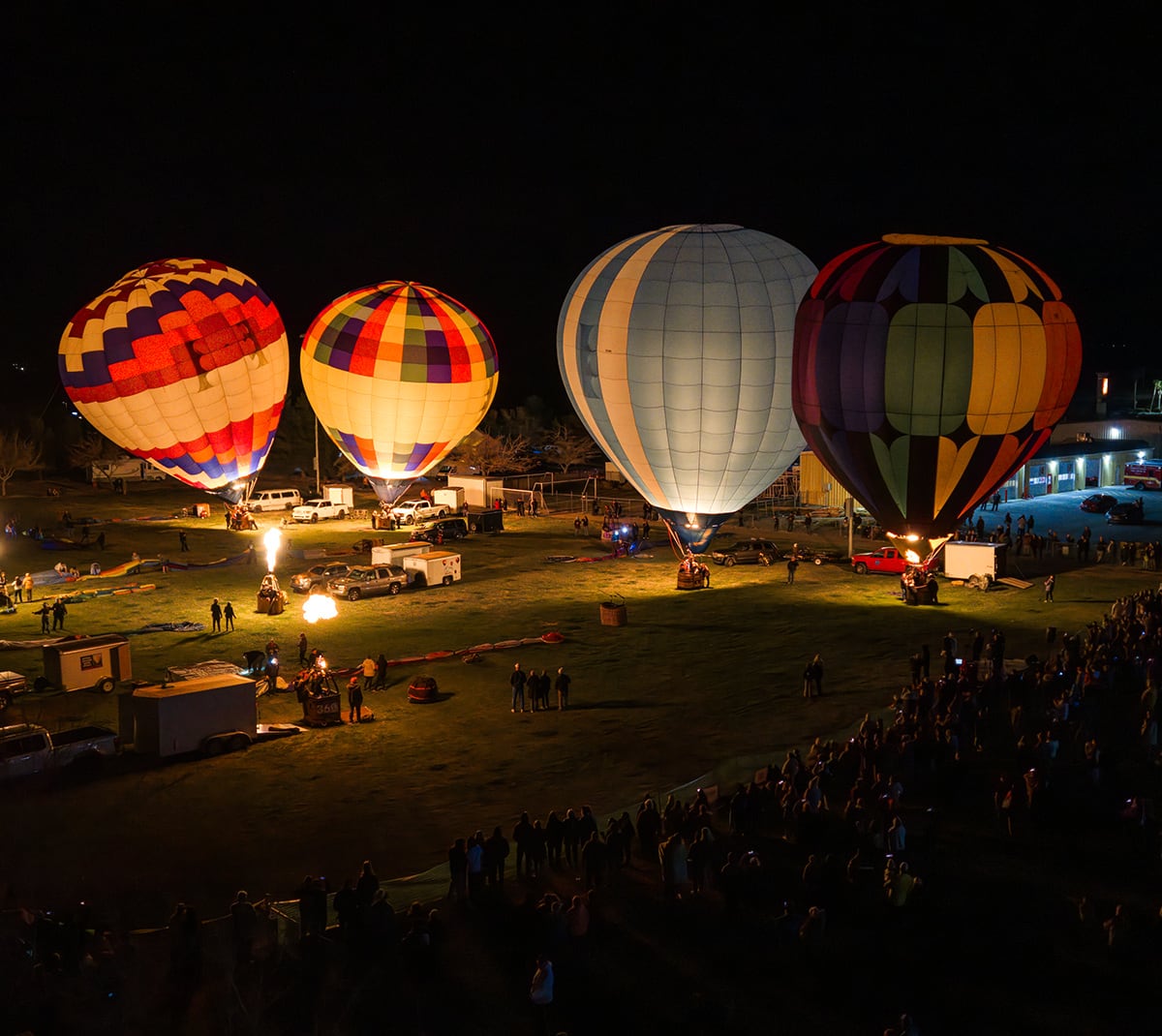 Four colorful hot air balloons glowing at night, surrounded by a crowd on a grassy field.