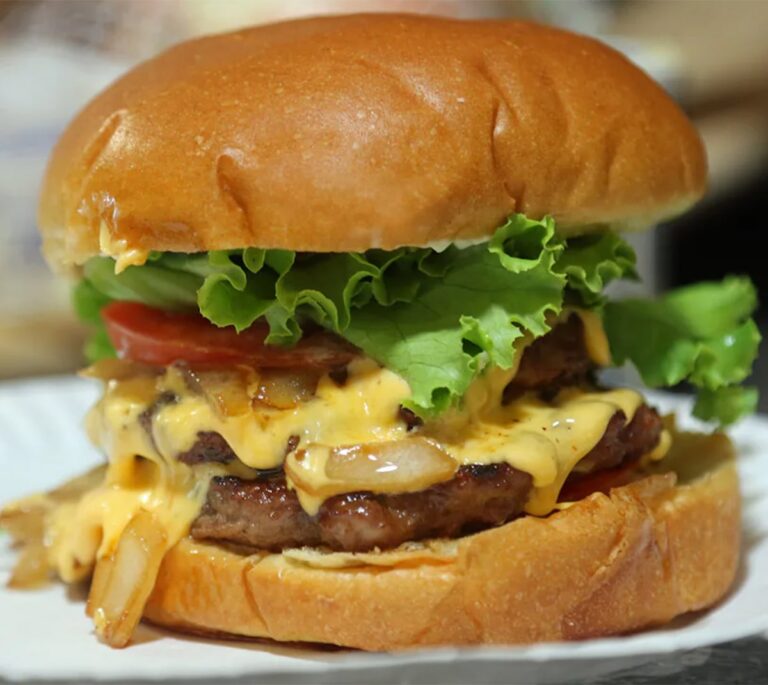A double cheeseburger with lettuce, tomato, melted cheese, onions, and a soft bun on a white plate.