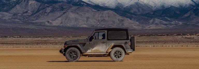 A muddy Jeep parked on a dry, flat desert with mountains in the background.
