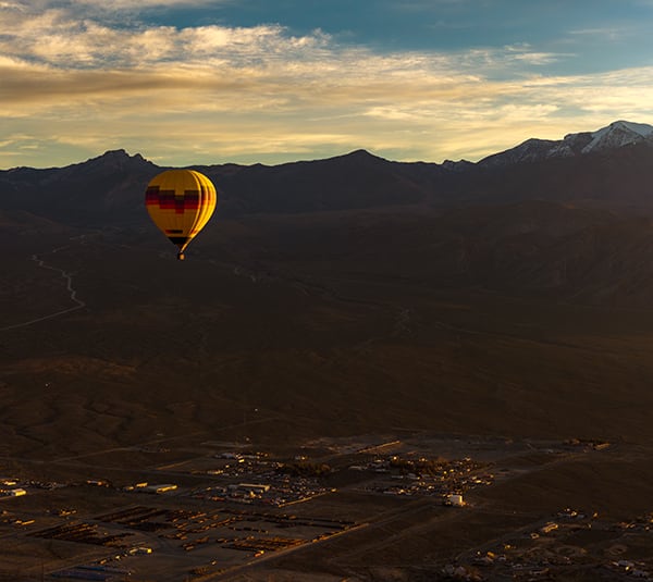 A yellow hot air balloon floats above a valley with mountains in the background at sunset.