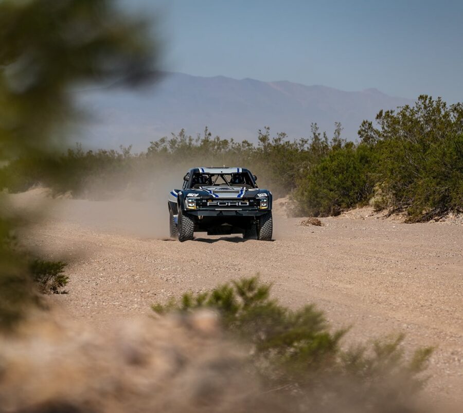 A black off-road truck drives on a dusty desert trail surrounded by bushes and distant mountains.