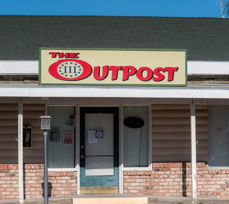 Storefront with a sign reading The Outpost above a door and windows, on a beige and brick building.