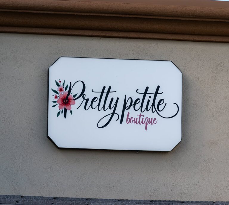 White sign with floral design reads Pretty Petite boutique in elegant script on a beige wall.