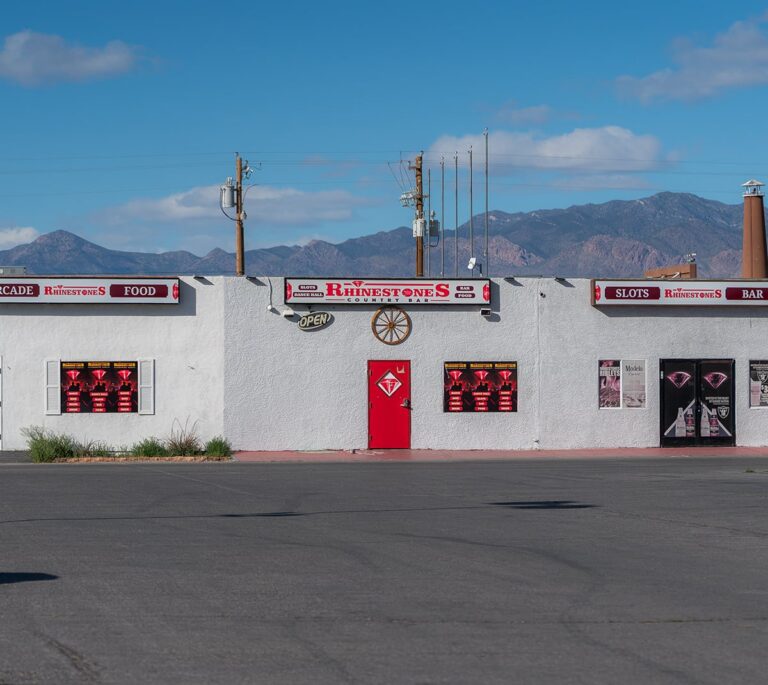 A white building with a red door and signs for Rhinestones bar, set against a mountain backdrop under a blue sky.