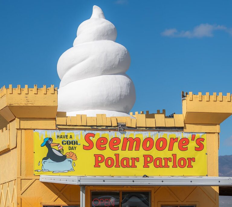 Yellow ice cream shop with a large white swirl sculpture on the roof and a playful penguin sign in front.