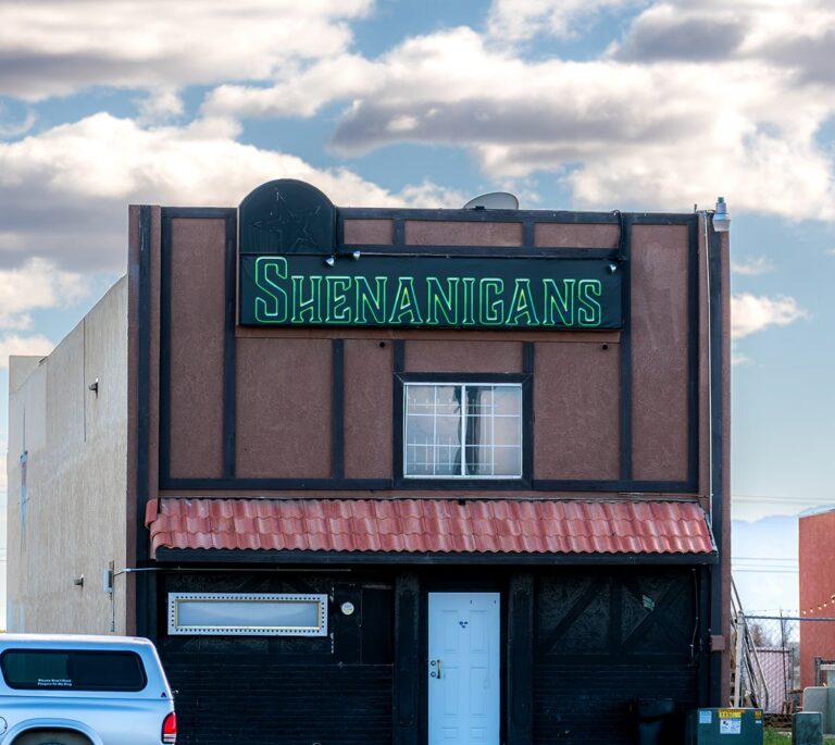 A small building with a green neon Shenanigans sign above the entrance under a cloudy sky.