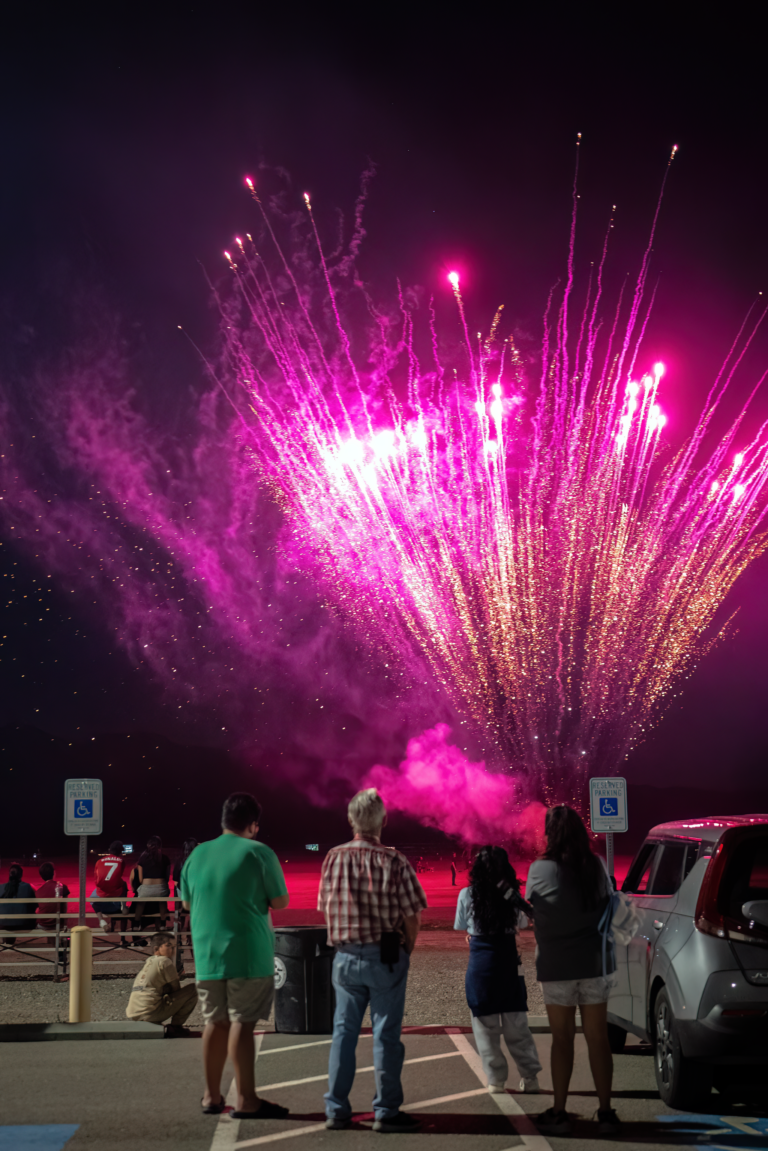 Four people watch vibrant pink fireworks lighting up the night sky in a parking lot.
