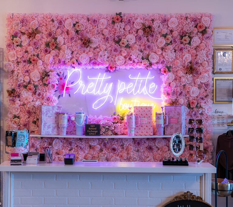 A boutique counter with pink floral wall, neon Pretty petite boutique sign, and various beauty products displayed.