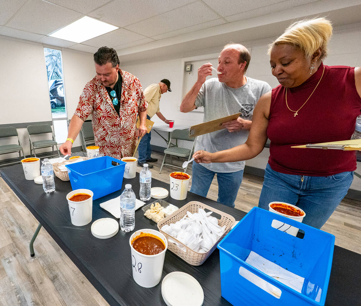 Three people taste and judge chili at a table with numbered bowls, water bottles, and utensils in a bright room.