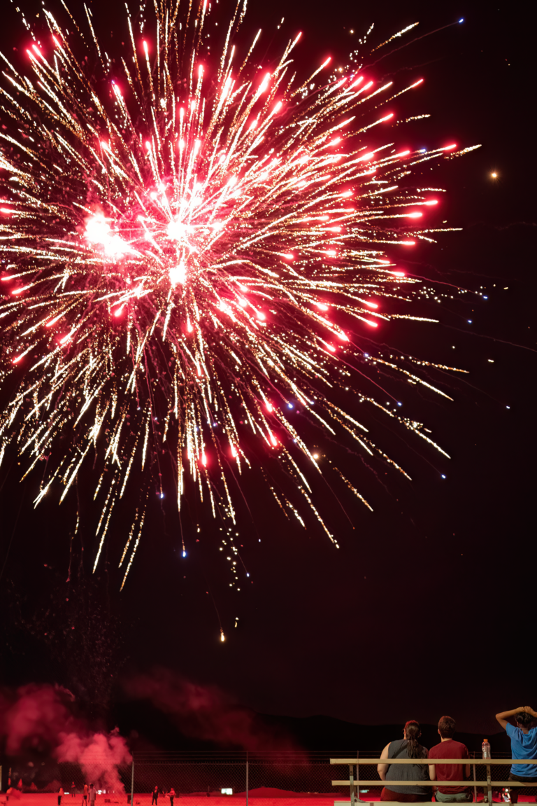 Large red fireworks burst in the night sky above people watching from bleachers.