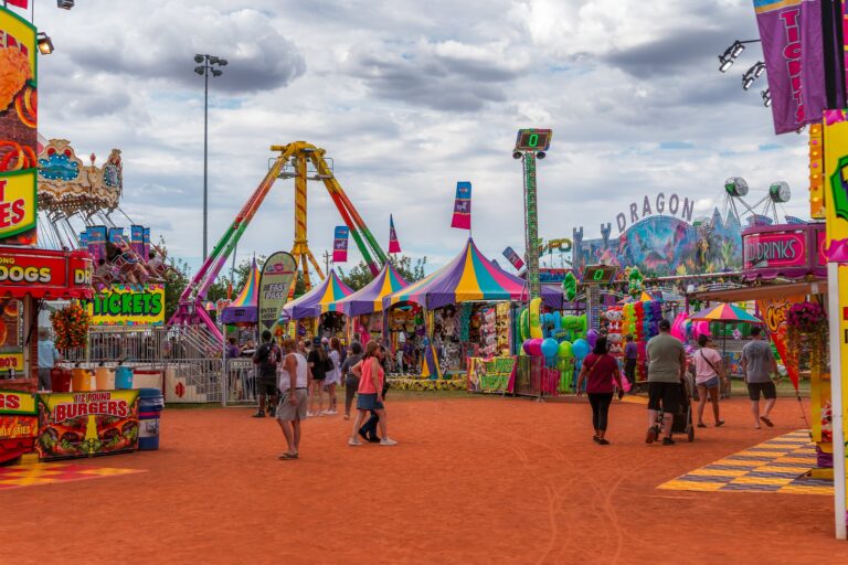 People walking through a colorful carnival with rides, games, food stands, and bright banners under a cloudy sky.