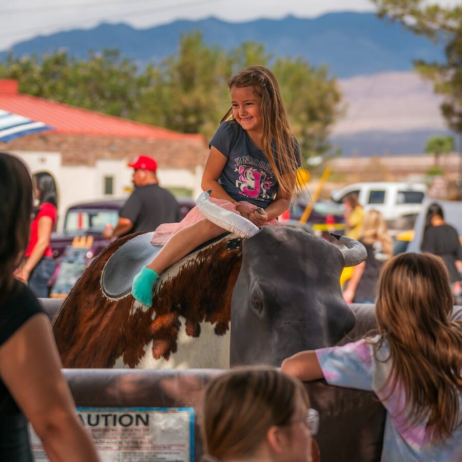 Smiling girl rides a mechanical bull at an outdoor event, with people watching and mountains in the background.