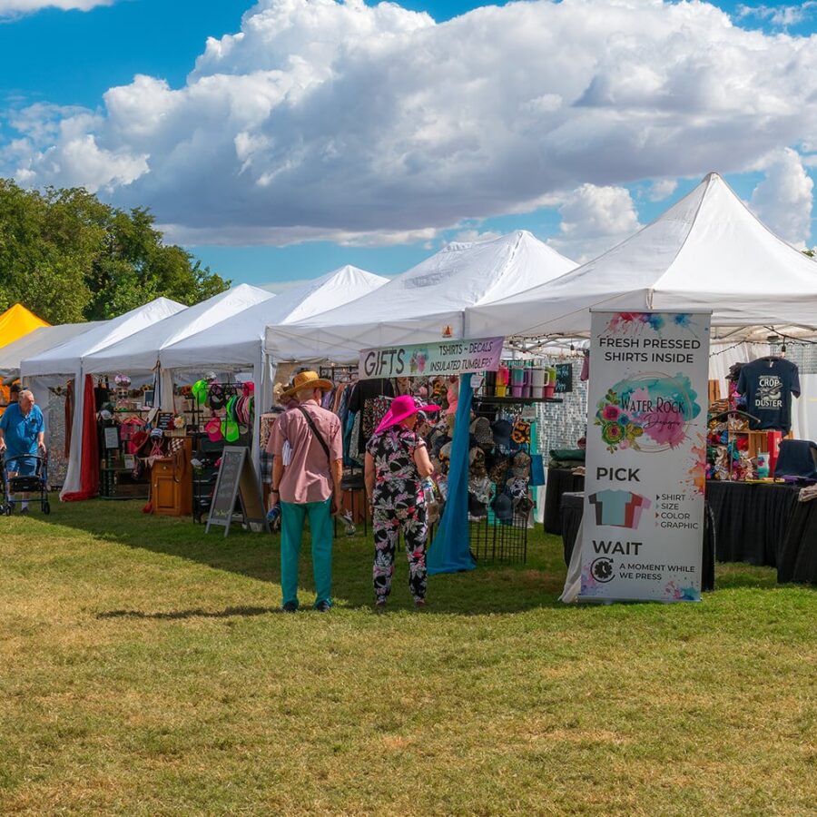 Outdoor market with white tents, people browsing gifts, shirts, and accessories on a grassy lawn under a blue sky.