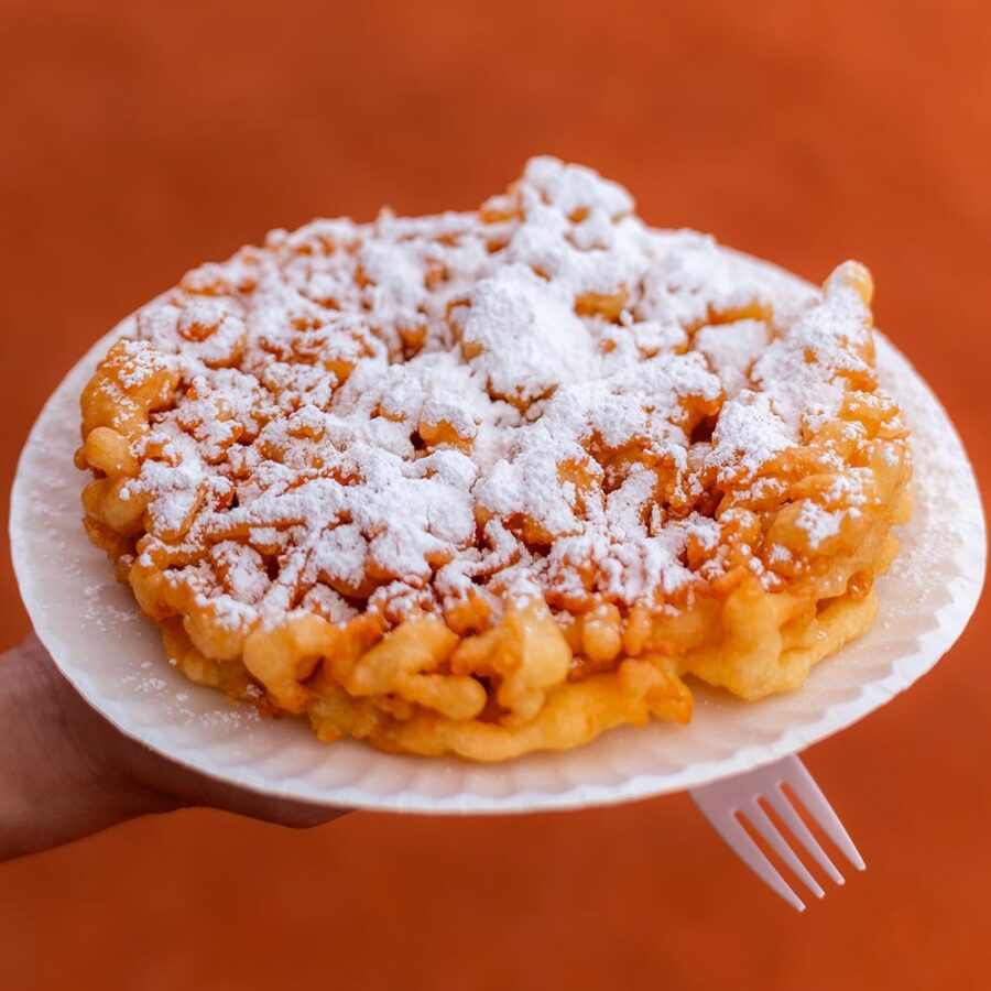 A hand holds a funnel cake topped with powdered sugar on a white plate, with an orange background.