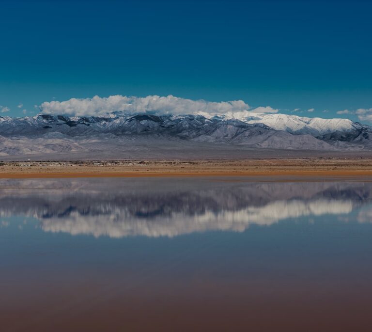 Snow-capped mountains reflected in a still lake under a clear blue sky with some clouds.