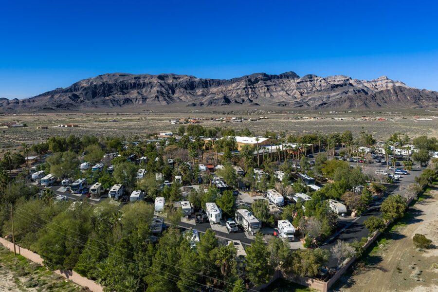 Aerial view of an RV park surrounded by trees, with mountains and desert landscape in the background.