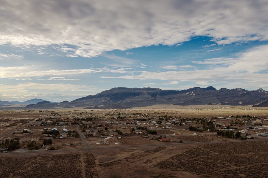 A small desert town with scattered houses, set against distant mountains under a partly cloudy sky.