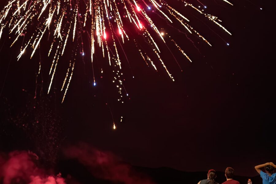 People watch colorful fireworks exploding in the night sky, with smoke rising in the foreground.