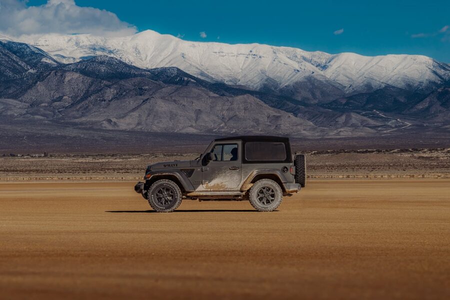 A black Jeep Wrangler on a flat, sandy plain with snow-capped mountains in the background.