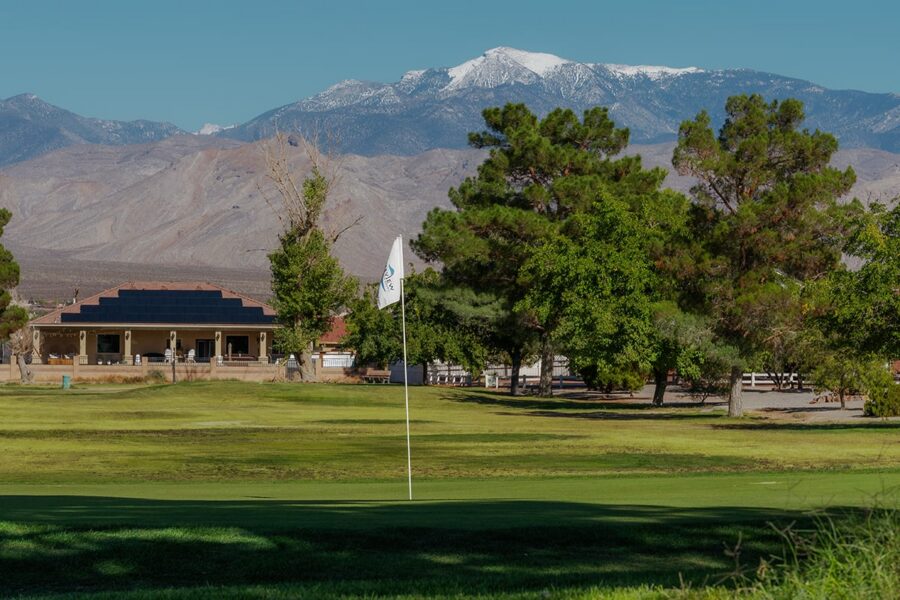 A golf course green with a flag, trees, a clubhouse, and mountains in the background under a clear sky.