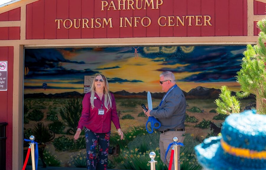 Two people stand by a ribbon at Pahrump Tourism Info Center, one holding large scissors for a ribbon-cutting.