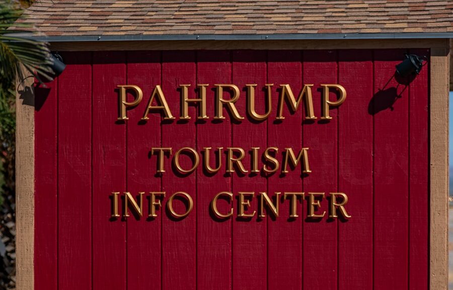 Red wall with gold letters reading Pahrump Tourism Info Center next to a large rock and some trees.