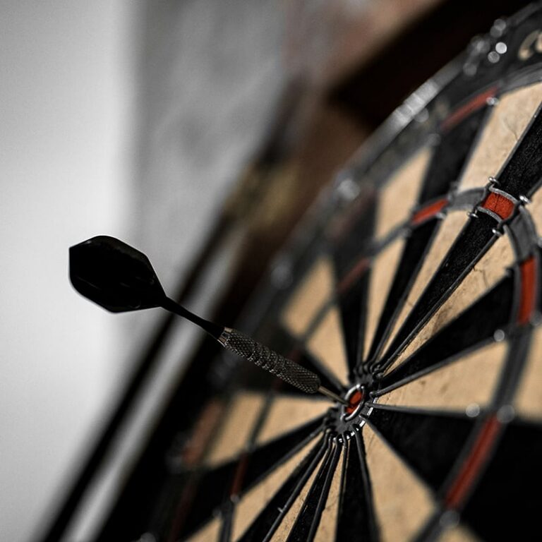 A dart sticks perfectly in the bullseye of a dartboard, shown in close-up with a blurred background.