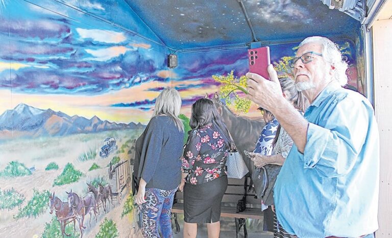 Man takes a photo with his phone as others view a colorful mural of a desert scene with mountains and horses.