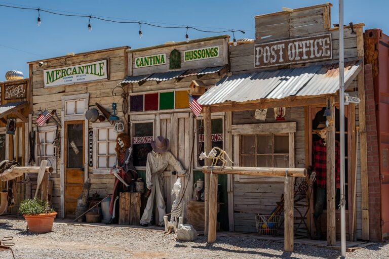 Wooden Old West-style storefronts with signs for mercantile, cantina, and post office on a sunny day.