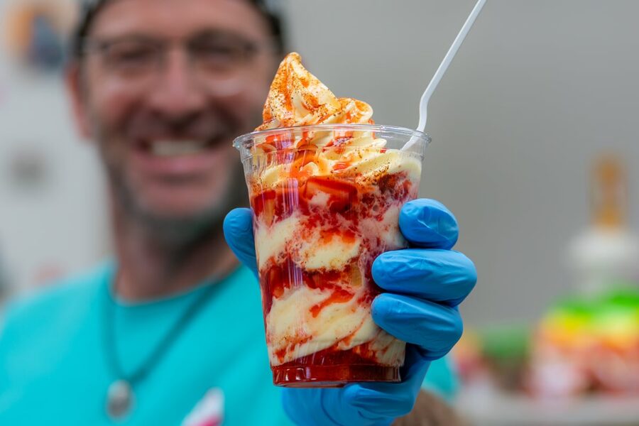 A person in blue gloves holds up a strawberry sundae with whipped cream and a spoon.