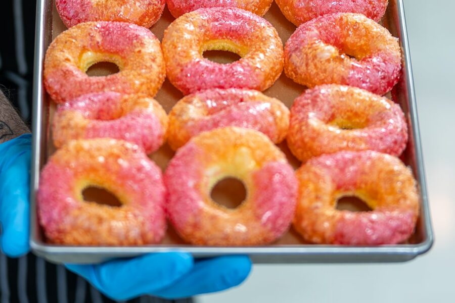 Tray of sugar-coated donuts, half pink and half orange, held by someone wearing blue gloves.