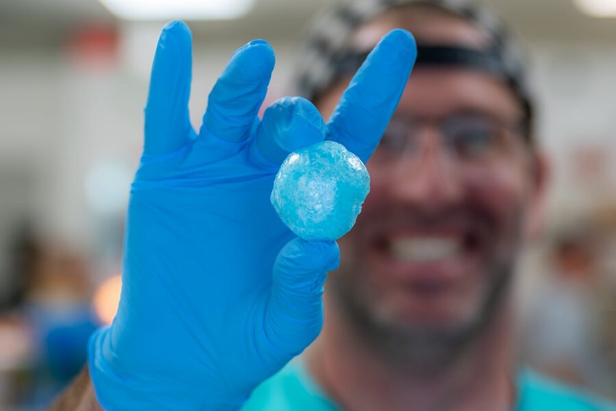 Person in blue gloves holds up a small, round blue object, smiling in a blurry background.