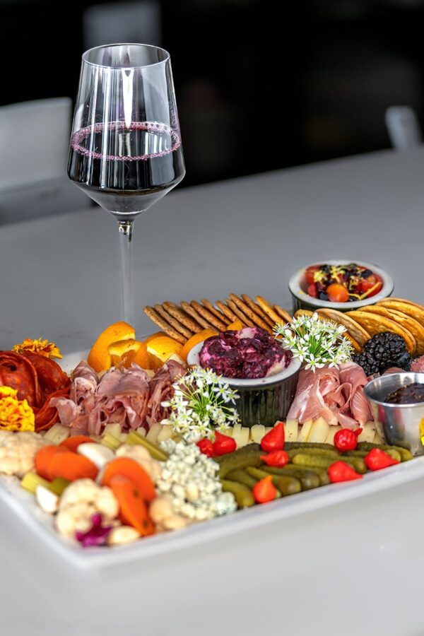 Charcuterie board with meats, cheese, fruit, crackers, and a glass of red wine on a white table.