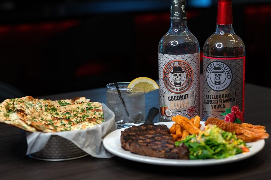 Grilled steak, fries, salad, flatbread, a cocktail, and two bottles of flavored liquor on a dark table.