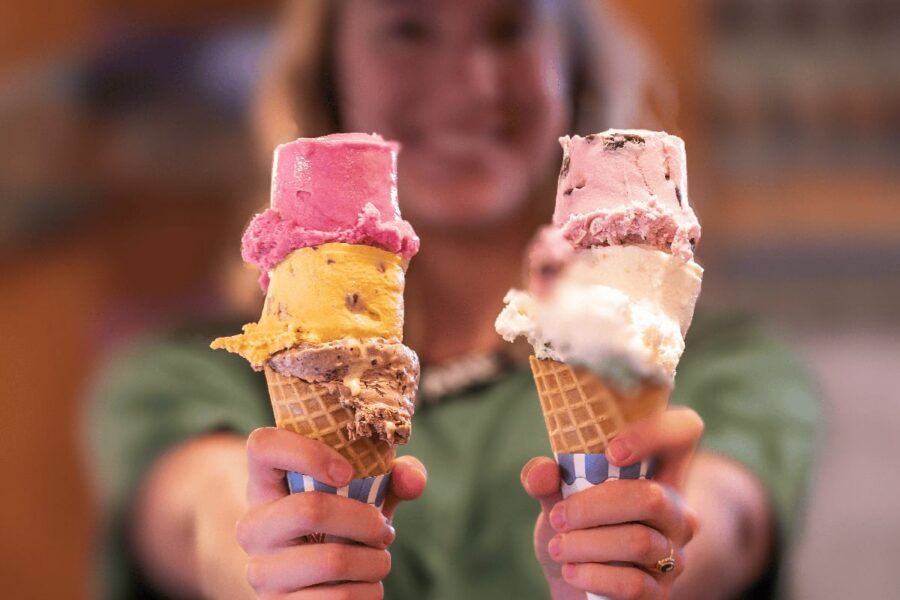 Person holding two ice cream cones, each stacked with three colorful scoops, blurred background.