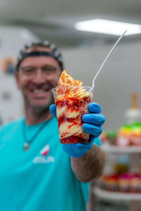 Person in blue gloves holds out a strawberry sundae in a clear cup with a white spoon.