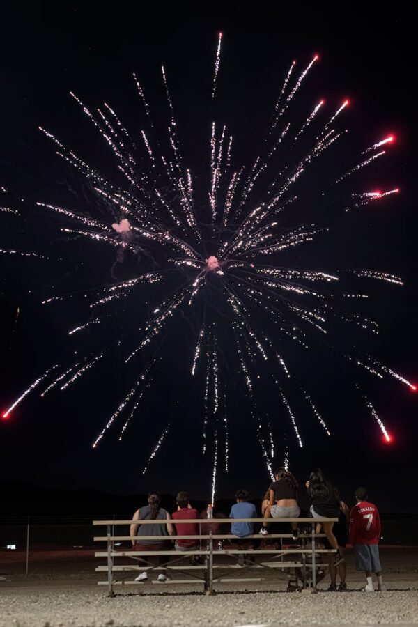 People sitting on bleachers at night watch large fireworks bursting in the dark sky above them.