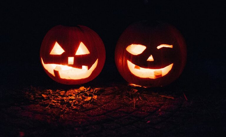 Two carved jack-o-lantern pumpkins glow in the dark, surrounded by fallen leaves on the ground.