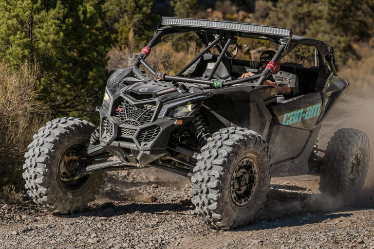 A black Can-Am off-road vehicle driving on a rocky dirt trail surrounded by trees and bushes.
